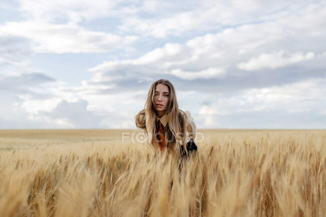 Young female with wavy hair looking at camera bending forward in countryside field under cloudy sky on blurred background — Stock Photo