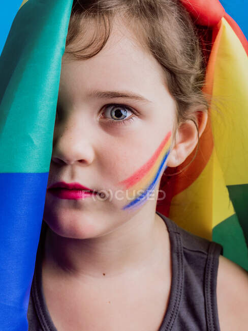 Cute girl with red lips and rainbow flag covering half of head looking at camera on blue background — Stock Photo