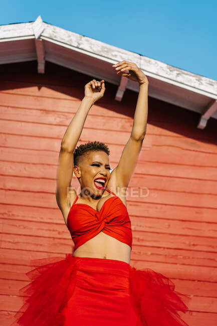 Trendy African American female with red lips and short hair dancing with raised arms while shouting against house wall — Stock Photo