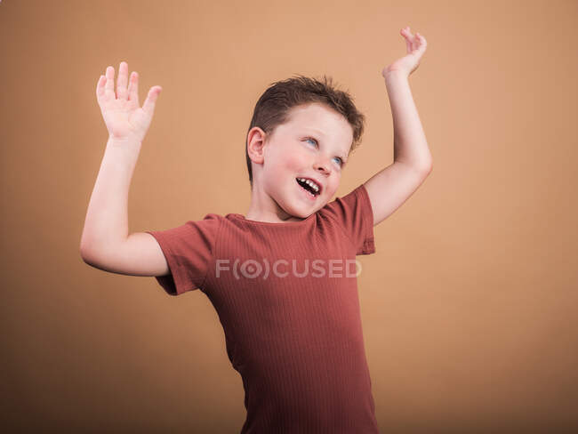 Задоволена дитина в повсякденному одязі з коричневим волоссям дивиться з зубною посмішкою і нахиленою головою, поки він підняв руки — стокове фото