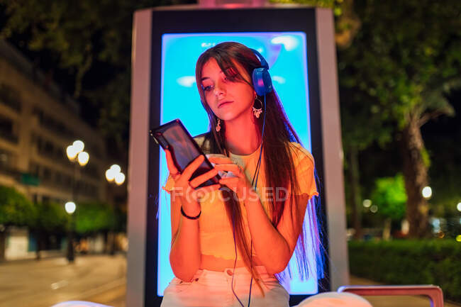 Interested female with beverage chatting on cellphone while listening to music from headset in night city — Stock Photo