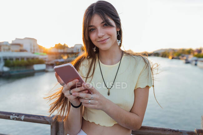 Sincere young female in pendant with cell phone over urban river in evening while looking at camera — Stock Photo