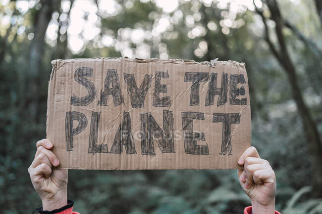 Criança étnica irreconhecível cortada mostrando o título de Save The Planet na peça de papelão enquanto olha para a câmera na floresta — Fotografia de Stock
