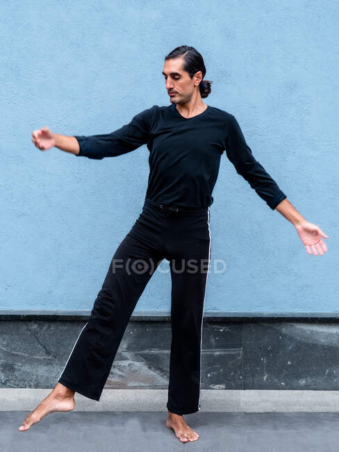 Focused handsome male dancer moving gracefully on city street against blue wall during rehearsal — Stock Photo