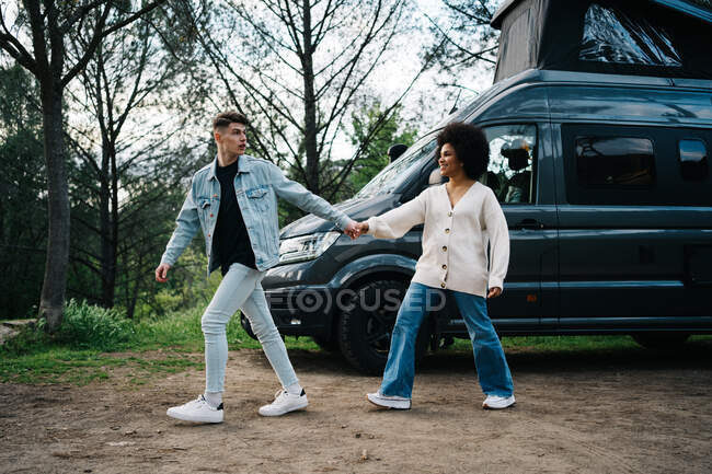 Sonriente mujer afroamericana con novio de moda cogido de la mano mientras pasea y mira hacia otro lado contra el campista - foto de stock
