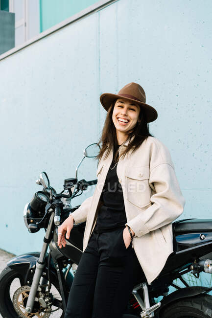Впевнена жінка-велосипедистка, що спирається на мотоцикл, припаркований на узбіччі міста і дивиться на камеру — стокове фото