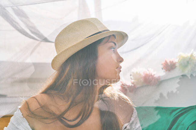 Хотя прозрачный занавес содержания молодой женщины в солнцезащитной шляпе охлаждения во дворе палатки в солнечный день и глядя в сторону — стоковое фото