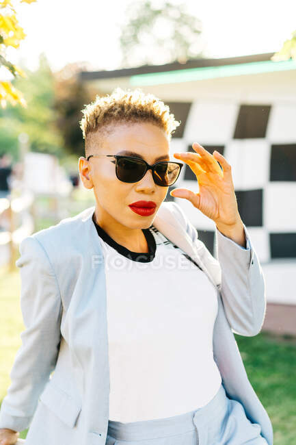 Portrait of African American female in trendy apparel with makeup and modern haircut looking at camera in park in back lit — Stock Photo