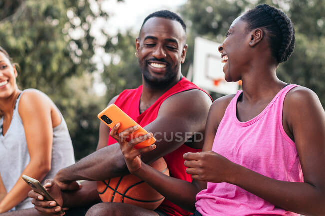 Delighted multiracial sportspeople browsing mobile phones and laughing on basketball sports ground in city — Stock Photo