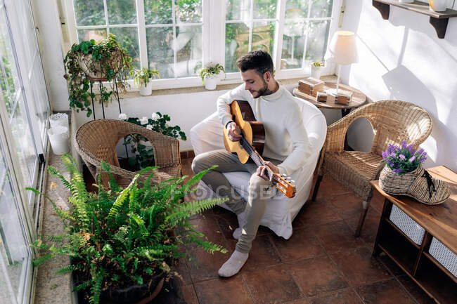 Созерцательный мужчина-музыкант с татуировками, играющий на классической гитаре, сидя в кресле и глядя в окно в доме — стоковое фото