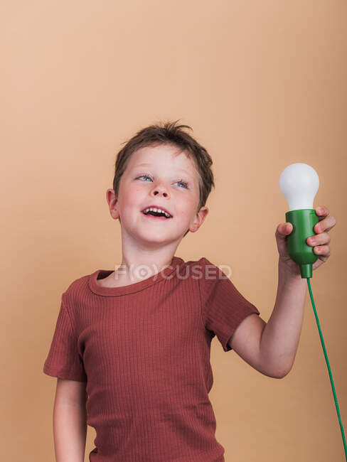 Pondering child in t shirt with plastic light bulb representing idea concept looking up on beige background — Stock Photo