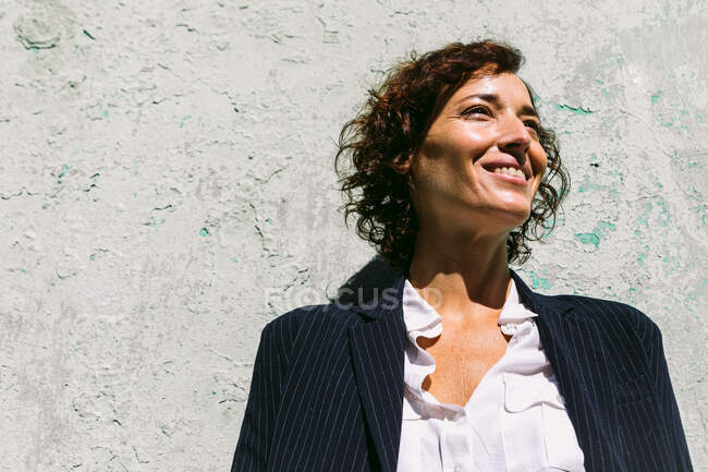 Счастливая женщина в элегантной одежде стоит солнечный свет на потрепанной стене и смотрит в камеру — стоковое фото