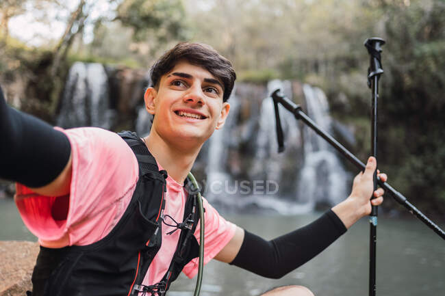 Escursionista maschio sorridente scattare auto sparato su smartphone mentre in piedi sullo sfondo della cascata e del lago nel bosco durante il trekking — Foto stock