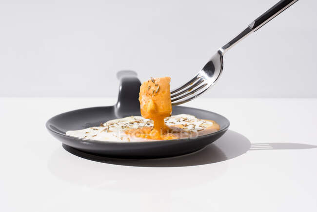 Garfo de metal com pedaço de pão fresco mergulhado em gema líquida de ovo frito servido em frigideira no fundo branco — Fotografia de Stock