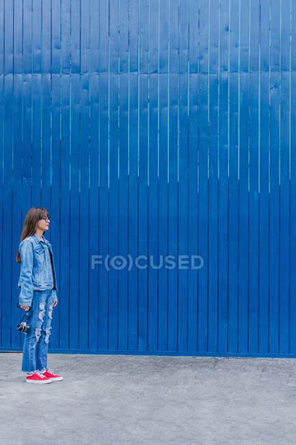 Vista laterale di fotografa donna in abiti di denim in piedi con fotocamera sullo sfondo della parete blu in strada — Foto stock