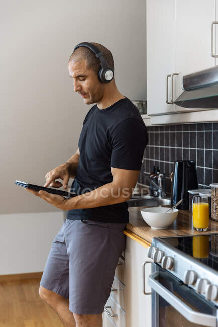 Seitenansicht des zufriedenen Mannes genießt Musik im Kopfhörer, während er morgens mit Tablet in der Küche steht — Stockfoto