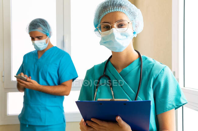 Жінка-лікар в медичній формі з доповіддю на буфері, стоячи в лікарні на тлі розмитого чоловічого лікаря — стокове фото
