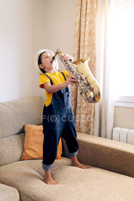 Criança descalça com olhos fechados tocando saxofone enquanto estava de pé no sofá em casa durante o dia — Fotografia de Stock