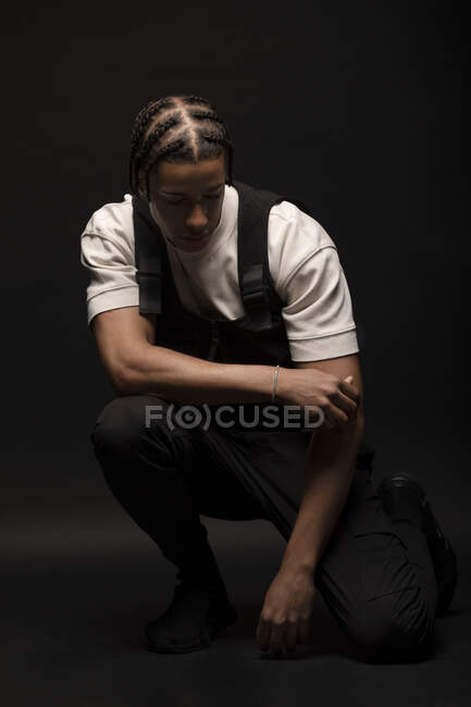 Side view of confident young ethnic hipster male model with braided hair wearing white shirt with black clothes looking down while crouched in dark studio — Stock Photo