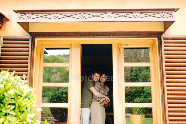 Vista do terraço do casal multirracial abraçando enquanto estava em pé no quarto perto de portas de casa de madeira e sorrindo — Fotografia de Stock