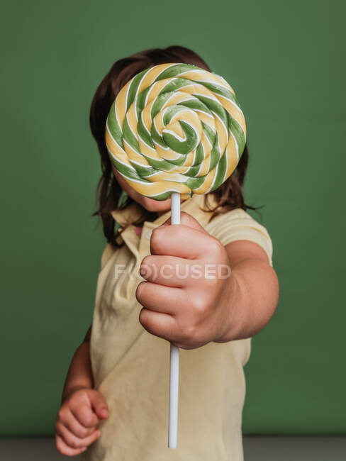 Anonymous kid reaching out hand with sweet swirl lollipop towards camera on green background in studio — Stock Photo
