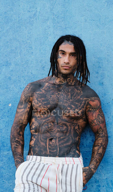 Hombre étnico de moda sin camisa con trenzas y tatuajes de pie cerca de la pared azul en la calle y mirando a la cámara - foto de stock