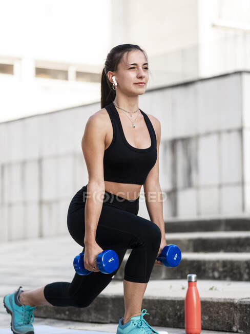 Jovem atleta feminina ouvindo música e olhando para o lado durante o exercício com halteres na rua da cidade — Fotografia de Stock