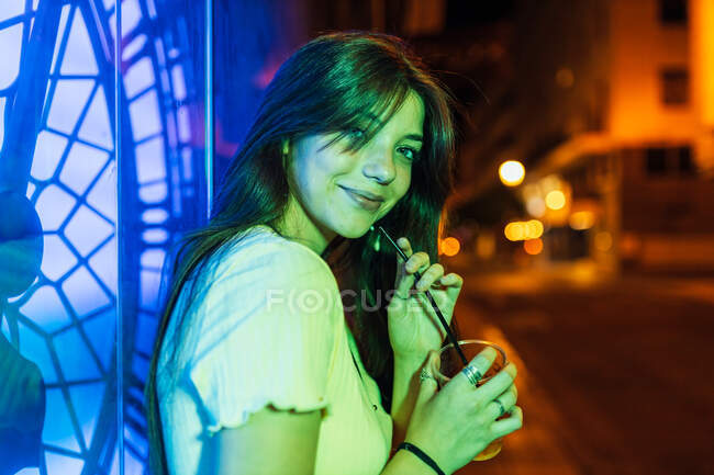 Contenida hembra joven con pelo largo y vaso de bebida refrescante mirando a la cámara al atardecer - foto de stock