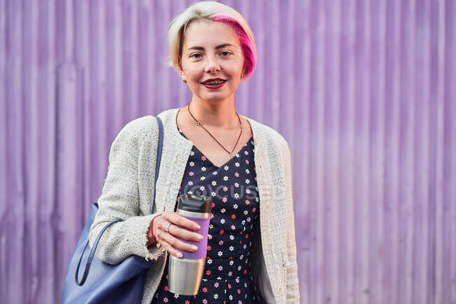 Mujer informal positiva con el pelo corto teñido de pie con bebida en copa ecológica en el fondo de la pared púrpura en la ciudad y mirando a la cámara - foto de stock