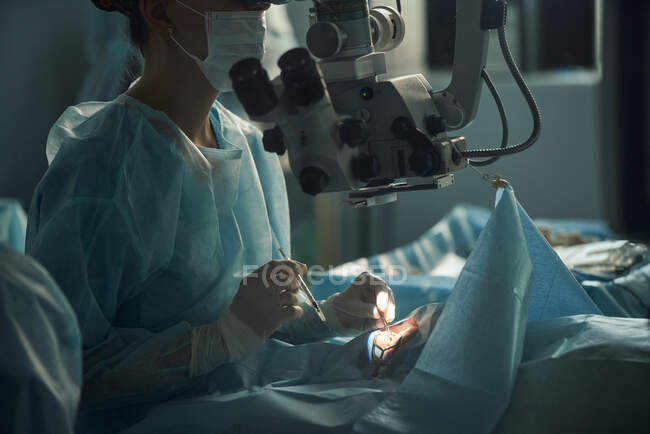 Medico donna adulto in maschera sterile e cappuccio medico ornamentale guardando attraverso il microscopio chirurgico contro il collega di coltura in ospedale — Foto stock