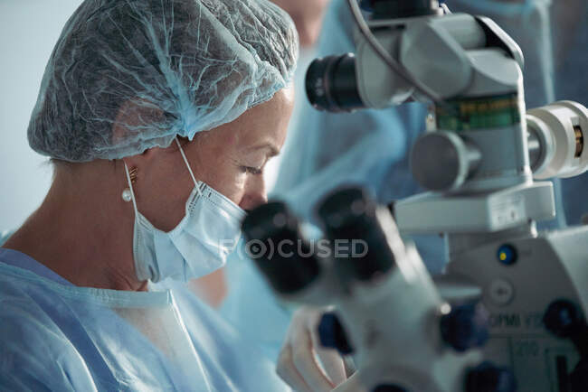 Зосереджений дорослий лікар у стерильній масці та орнаментальній медичній шапці, дивлячись через хірургічний мікроскоп проти співробітника врожаю в лікарні. — стокове фото