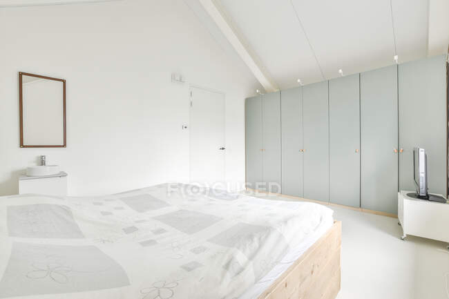 Luz ático dormitorio interior con paredes blancas amuebladas con cama y armario con TV en esquina en casa de estilo loft moderno - foto de stock