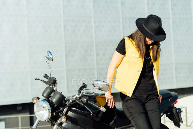 Anonymous female motorcyclist in hat leaning on modern motorbike parked on road in city on sunny day — Stock Photo