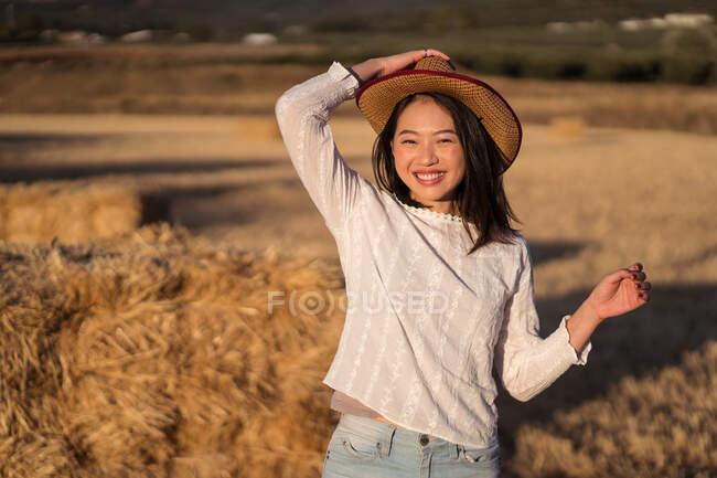 Contenuto femmina etnica con cappello di paglia in piedi vicino pila di fieno secco in campo e guardando la fotocamera — Foto stock