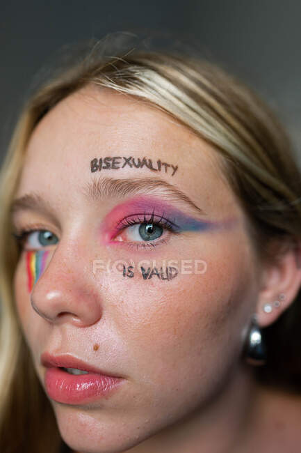 Lesbische Frau mit Inschrift im Gesicht Bisexualität ist gültig und Regenbogen-LGBT-Flagge blickt in die Kamera — Stockfoto
