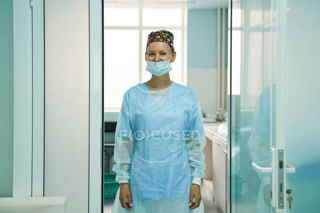Allegro medico femminile adulto in maschera sterile e cappuccio ornamentale guardando la fotocamera in ospedale — Foto stock