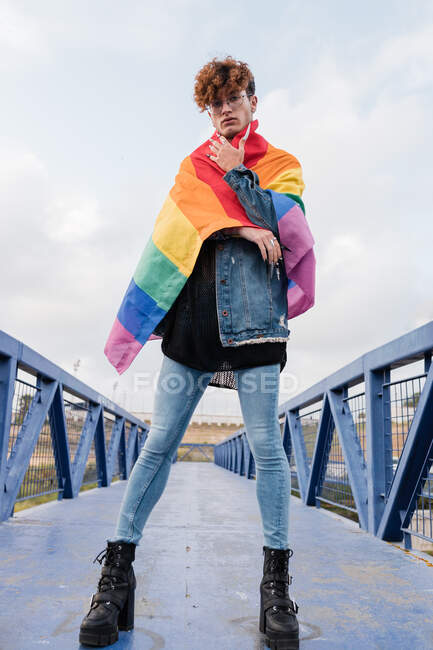 D'en bas d'un homme homosexuel sérieux debout avec drapeau arc-en-ciel sur le pont et regardant la caméra — Photo de stock