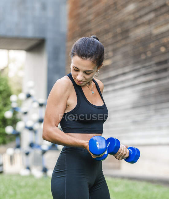 Determined female athlete doing exercise with dumbbells during fitness workout on city street in summer — Stock Photo