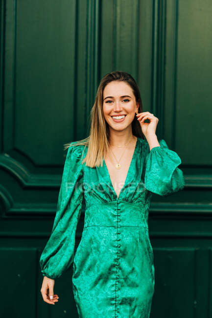 Fröhliche Frau in stilvollem grünem Kleid steht neben dekorativen Holztüren auf der Straße und blickt in die Kamera — Stockfoto