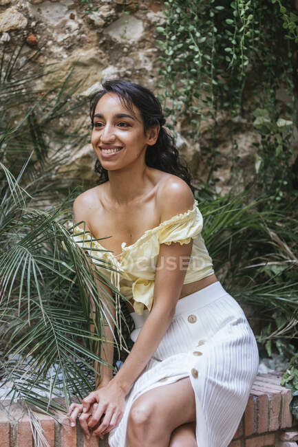 Deliciosa hembra étnica en traje de verano sentada en la frontera de piedra en un jardín exótico y mirando hacia otro lado - foto de stock