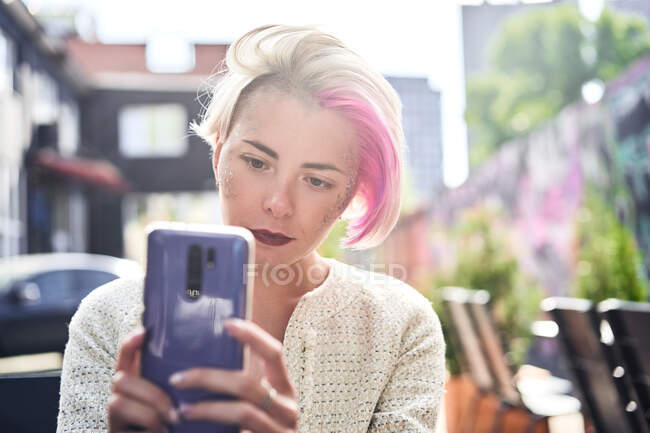 Mujer informal enfocada con el pelo corto teñido y con brillo en la cara que navega por el teléfono celular en la ciudad - foto de stock