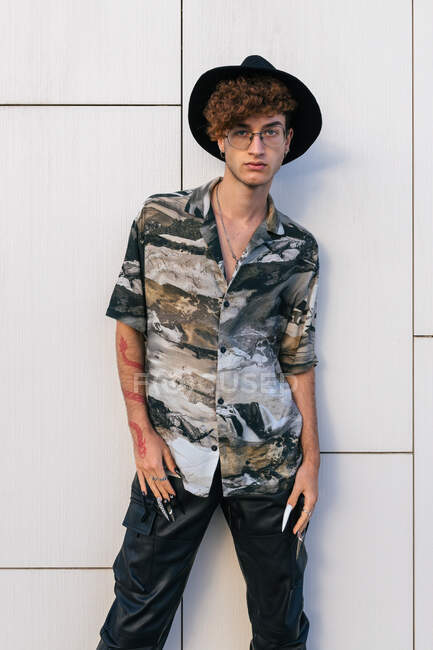 Jeune homme vaniteux en tenue élégante avec chapeau debout sur le mur carrelé regardant la caméra — Photo de stock