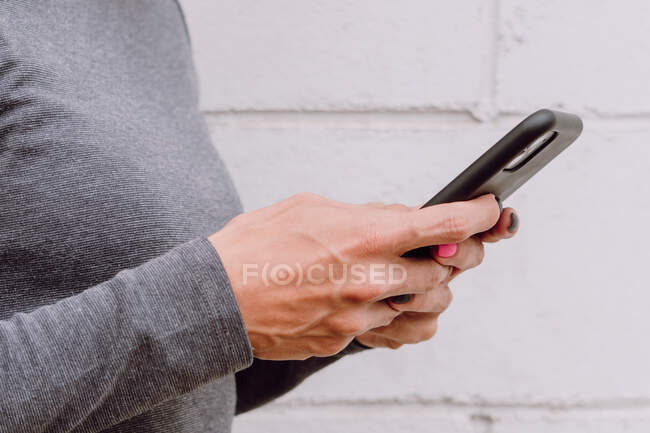 Mulher anônima em roupas casuais usando smartphone enquanto estava perto da parede branca na rua — Fotografia de Stock