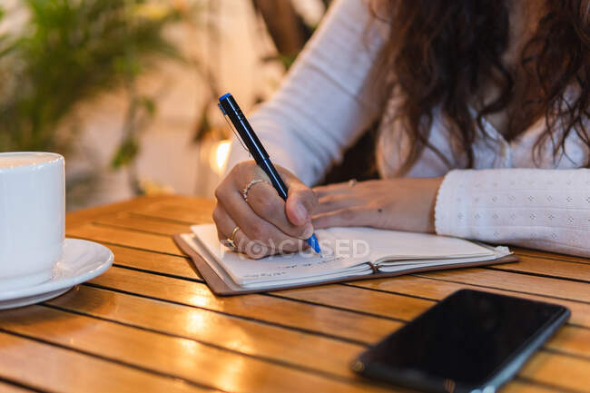 Cortada mujer de negocios étnica irreconocible en ropa casual tomando notas en planificador mientras se sienta a la mesa con una taza de café y teléfono inteligente en la acogedora cafetería con plantas verdes - foto de stock