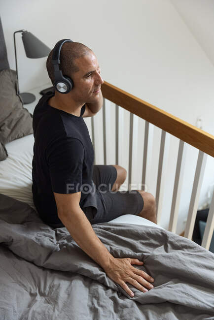 Зверху спокійний чоловік одягає навушники і слухає музику, сидячи в ліжку після пробудження в спальні і насолоджуючись ранком — стокове фото