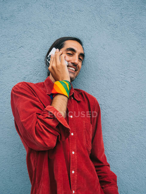 Зачарований етнічний гей-чоловік з веселкою банданою під рукою розмовляє на мобільному телефоні, стоячи біля сірої стіни в місті і дивлячись на камеру — стокове фото