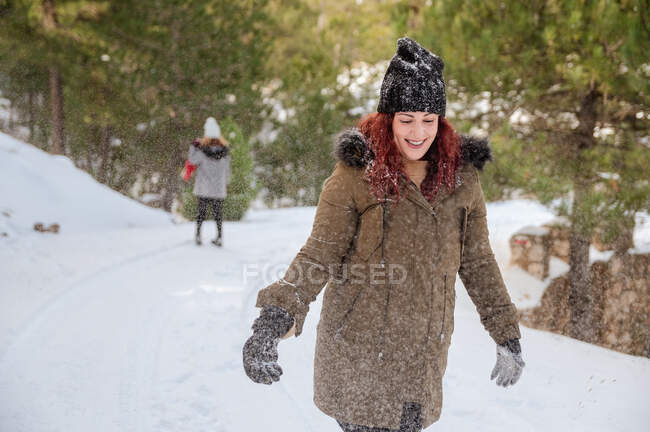 Весела жінка в верхньому одязі стоїть в зимовому лісі і кидає сніг, розважаючись — стокове фото