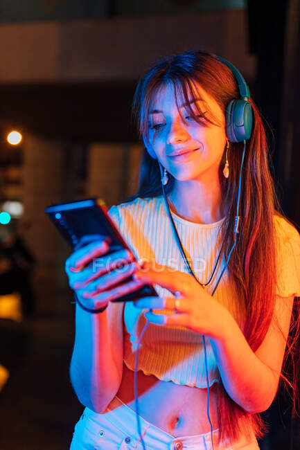 Cheerful young female with cellphone listening to song from headphones against colorful neon lights in evening town — Stock Photo