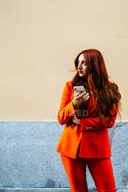 Stylish female with ginger hair and in bright orange suit standing in city street and using mobile phone while looking away — Stock Photo