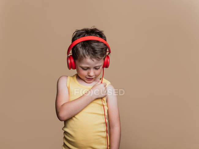Satisfeito menino pré-adolescente em fones de ouvido vermelhos ouvindo música em fundo marrom em estúdio — Fotografia de Stock